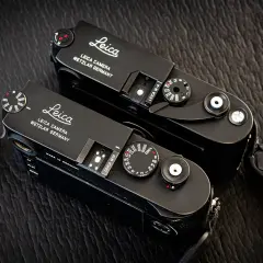 Two black Leica M cameras, a Leica M10 and a Leica M-A, sit side-by-side on a textured dark surface.