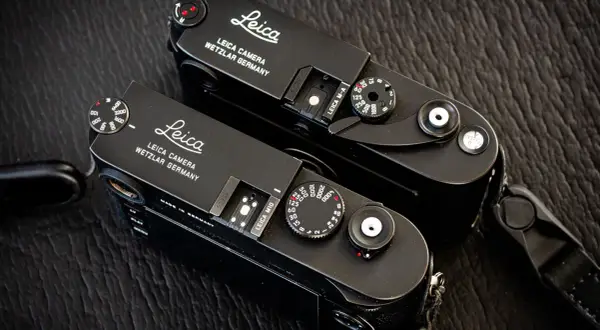 Two black Leica M cameras, a Leica M10 and a Leica M-A, sit side-by-side on a textured dark surface.