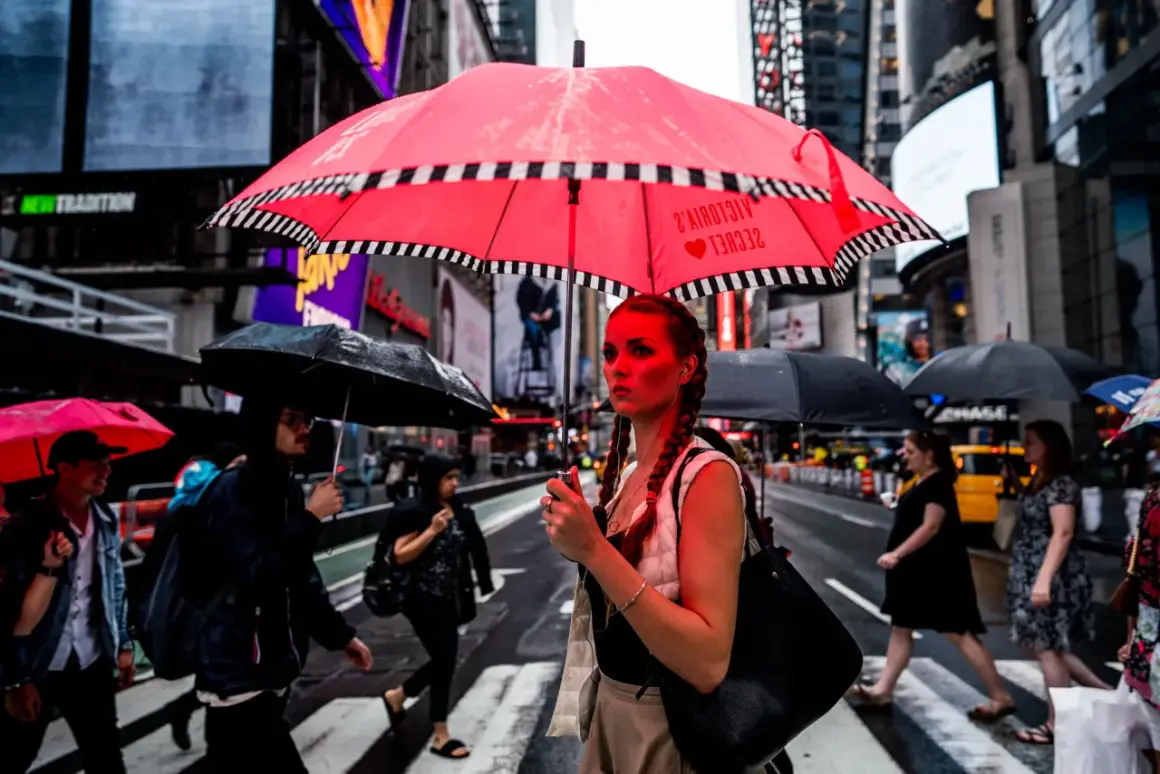 A woman with braided hair holds a bright red umbrella in this 24mm street photography shot. Rain falls on Times Square as people cross the street.