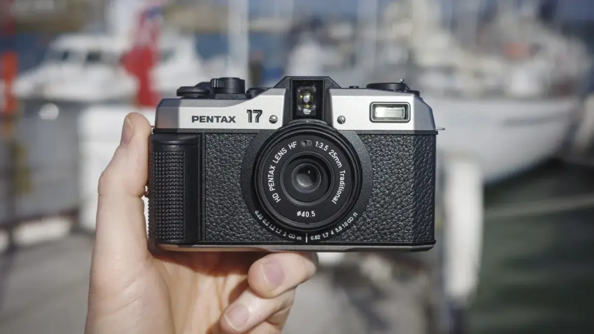 A hand holds the highest quality film camera, a silver and black Pentax 17, with a blurred marina in the background.