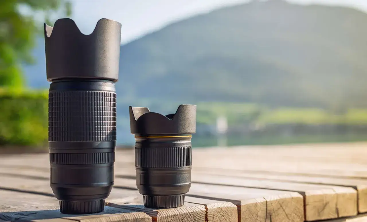 A tall telephoto lens stands beside a shorter, normal lens on a wooden surface, with a blurred mountain and lake in the background.