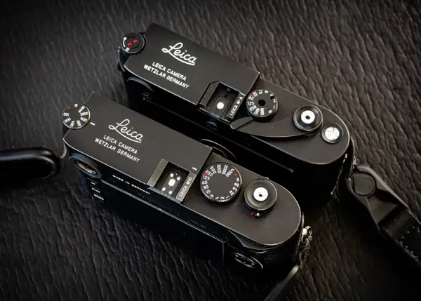 Two black Leica M cameras, a Leica M10 and a Leica M-A, sit side-by-side on a textured dark surface.
