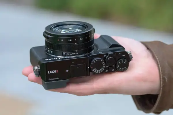 A hand holds a Panasonic Lumix DMC-LX100K camera, showcasing its compact design and prominent lens.